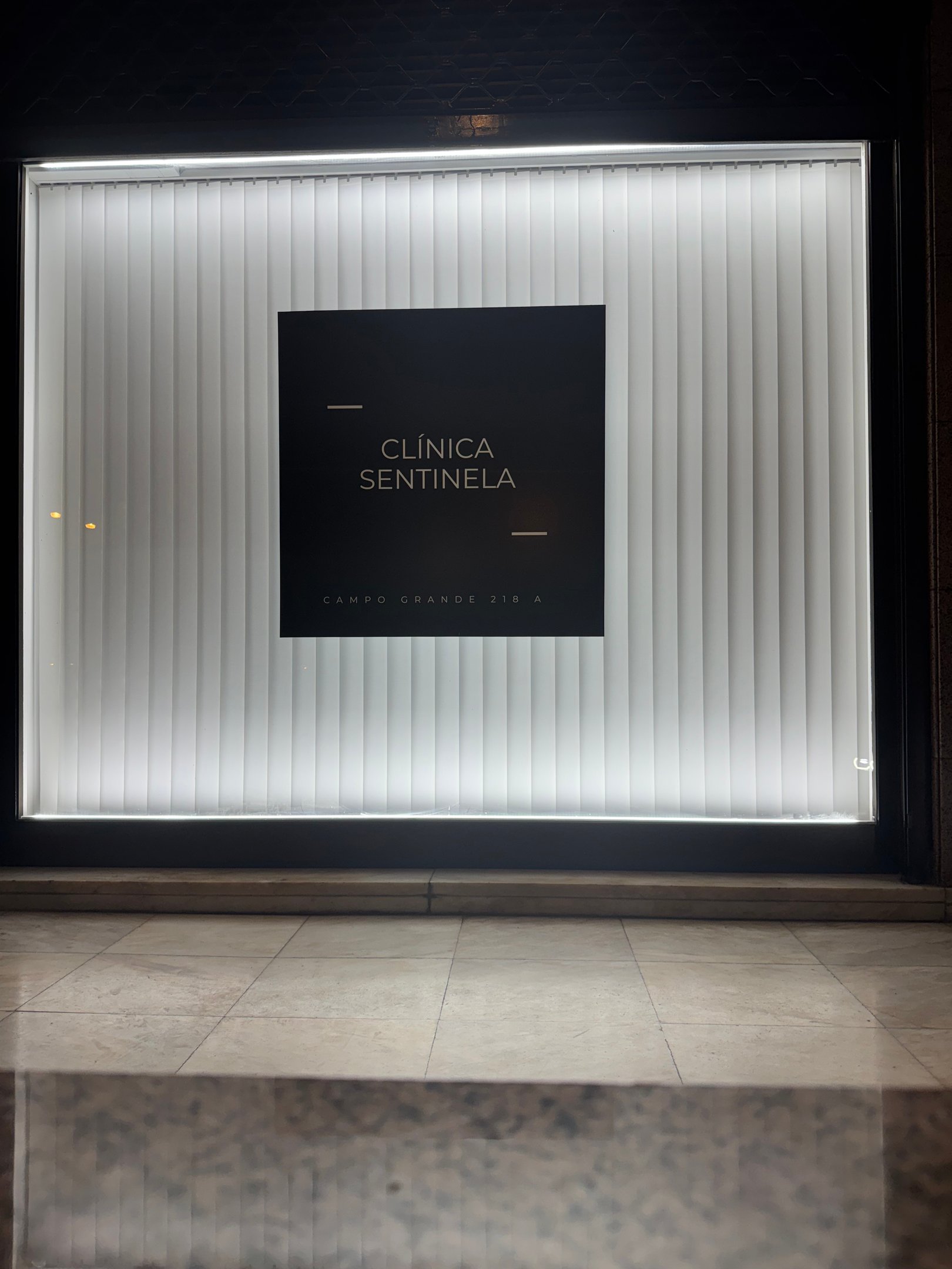 Illuminated Clínica Sentinela storefront window with black logo signage and white vertical blinds.