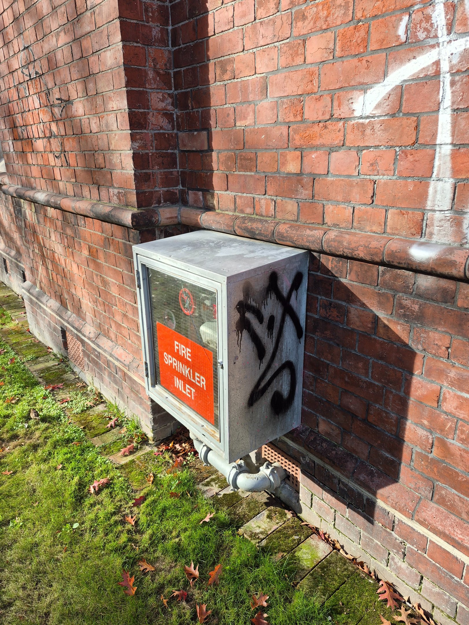 A metal fire sprinkler inlet box with graffiti mounted on an old red brick wall outdoors.