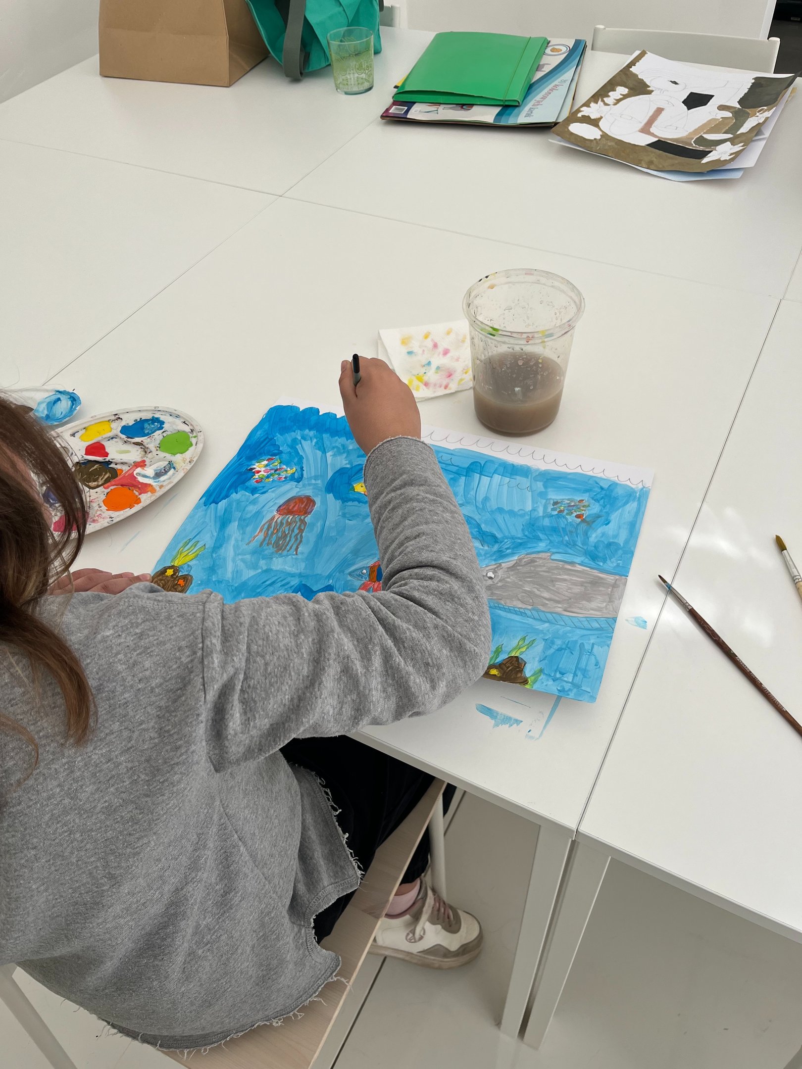 A kid at a desk painting a blue ocean scene with jellyfish and sea creatures.