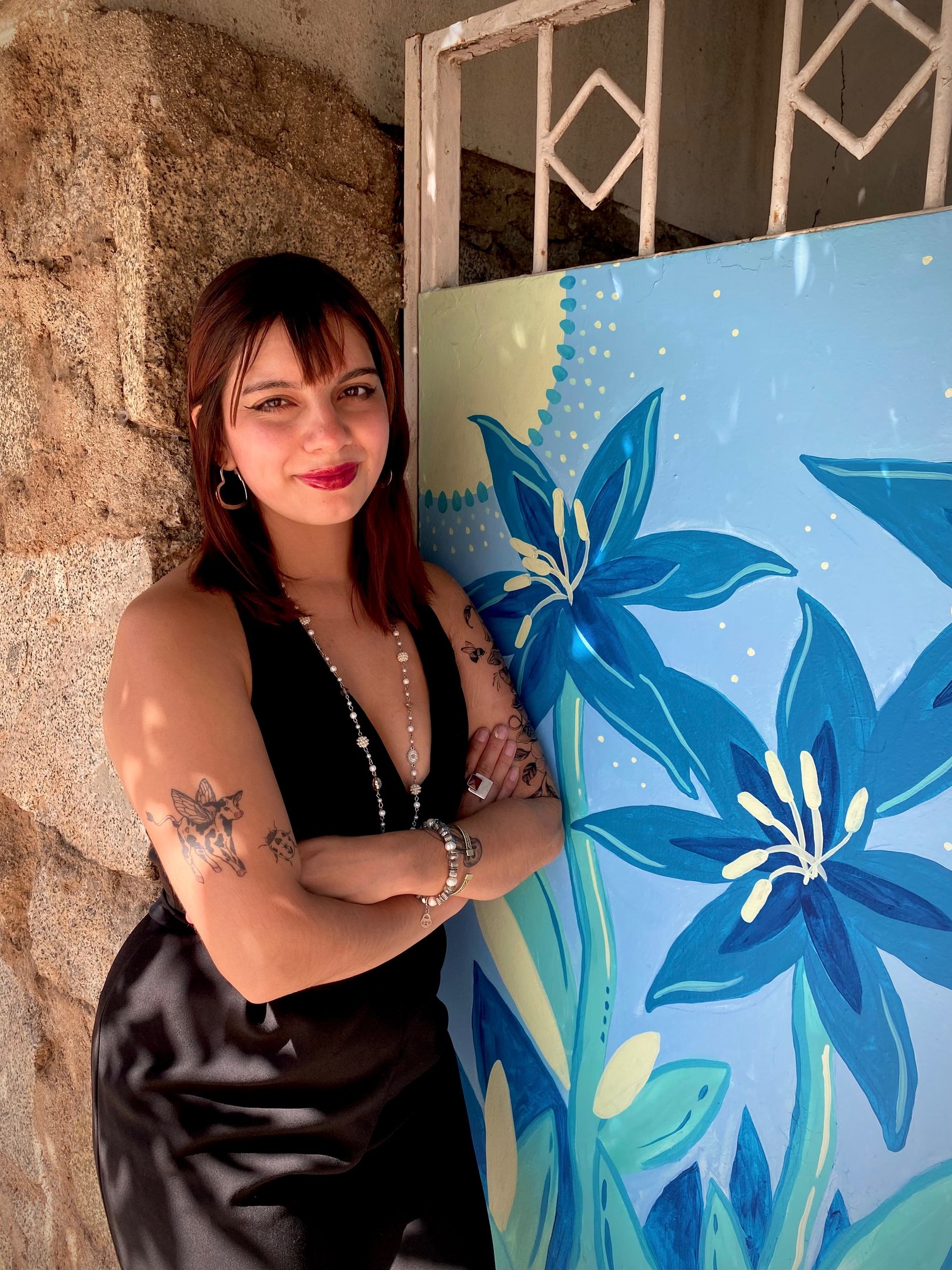 Young woman with tattoos posing in a black dress next to a blue floral mural.