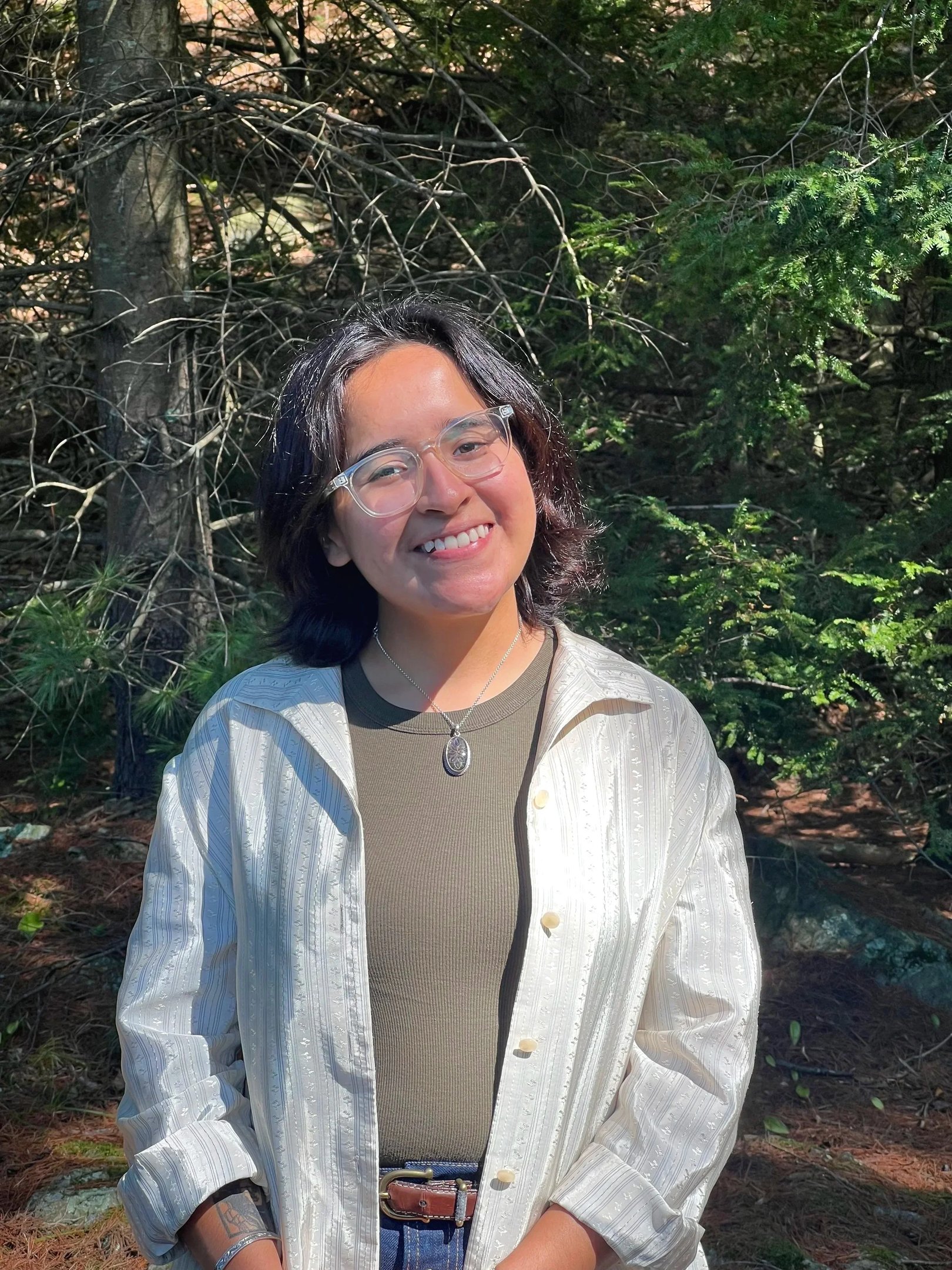 Smiling woman with glasses wearing a white shirt and olive top standing in a sunny evergreen forest.