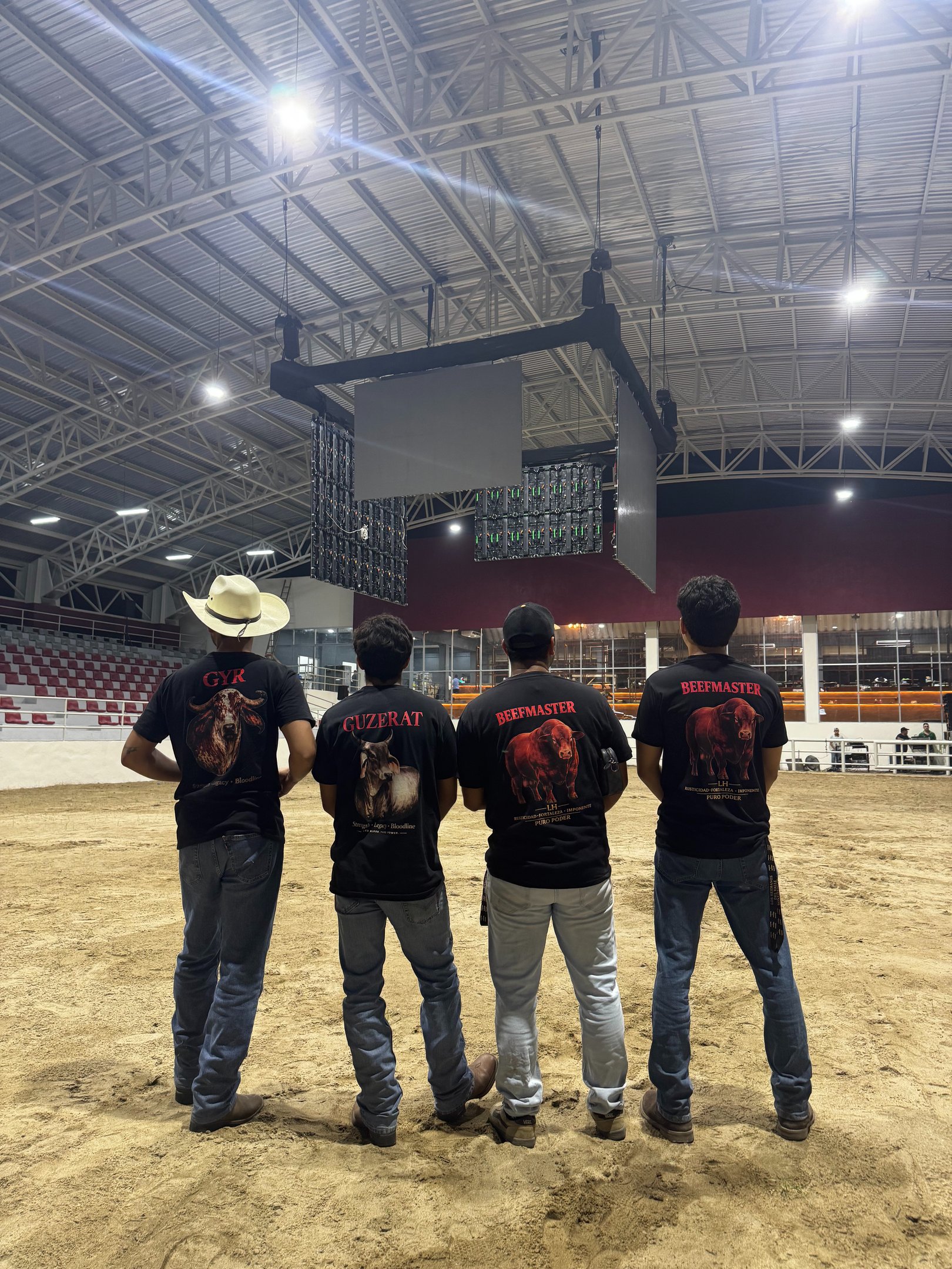 Four men in cattle breed t-shirts standing in a rodeo arena under indoor stadium lights.