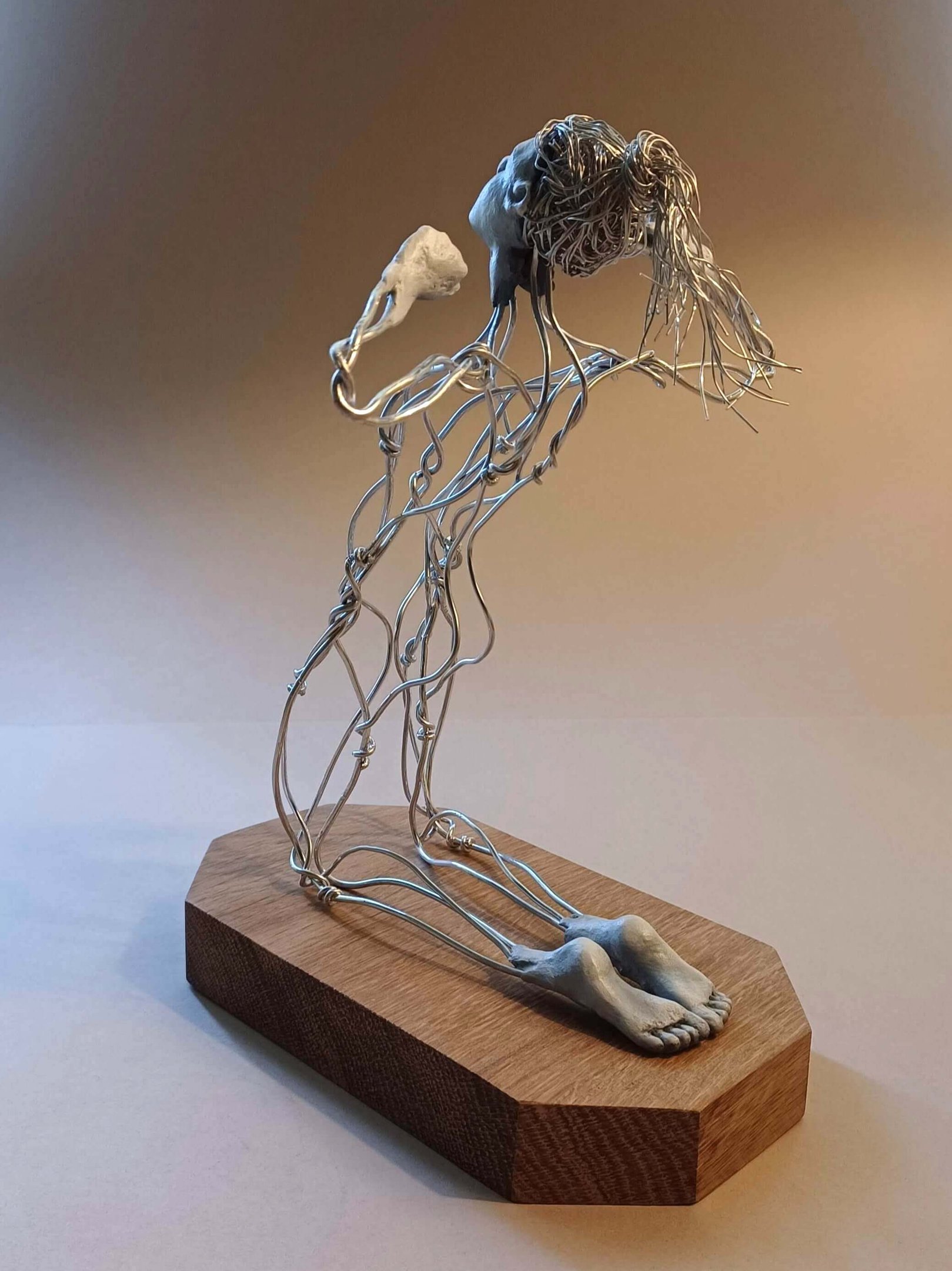 A wire sculpture of a complete kneeling female figure with clay hands, face and feet, arms raised