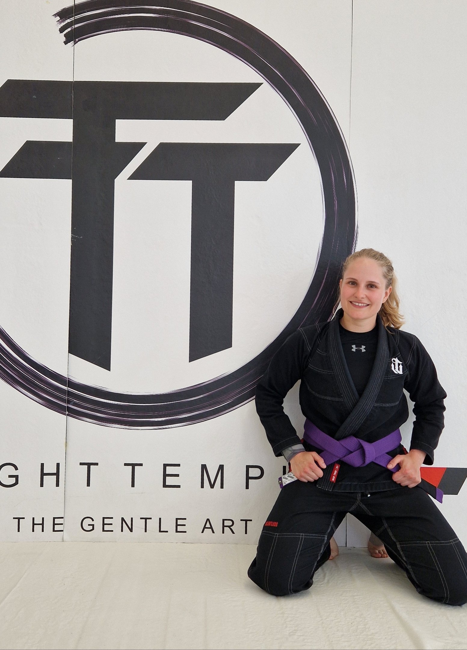 A female martial artist in a black BJJ gi and purple belt kneeling in front of a gym logo.