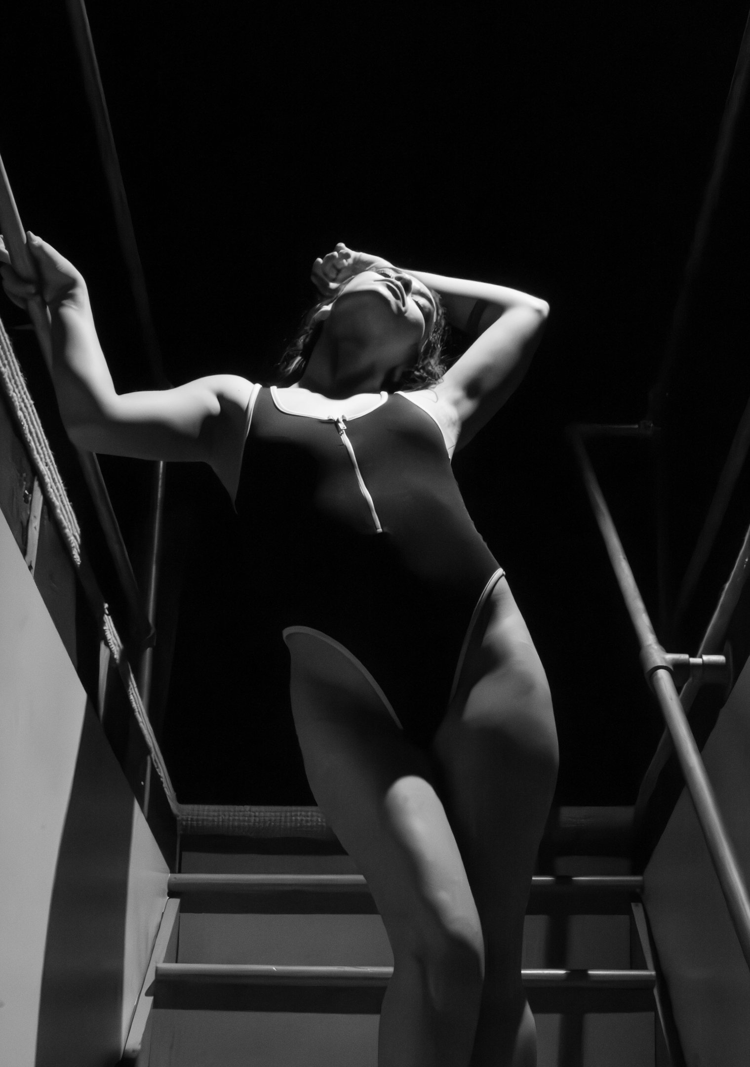 Black and white portrait of a woman in a one-piece swimsuit posing on stairs in dramatic lighting.