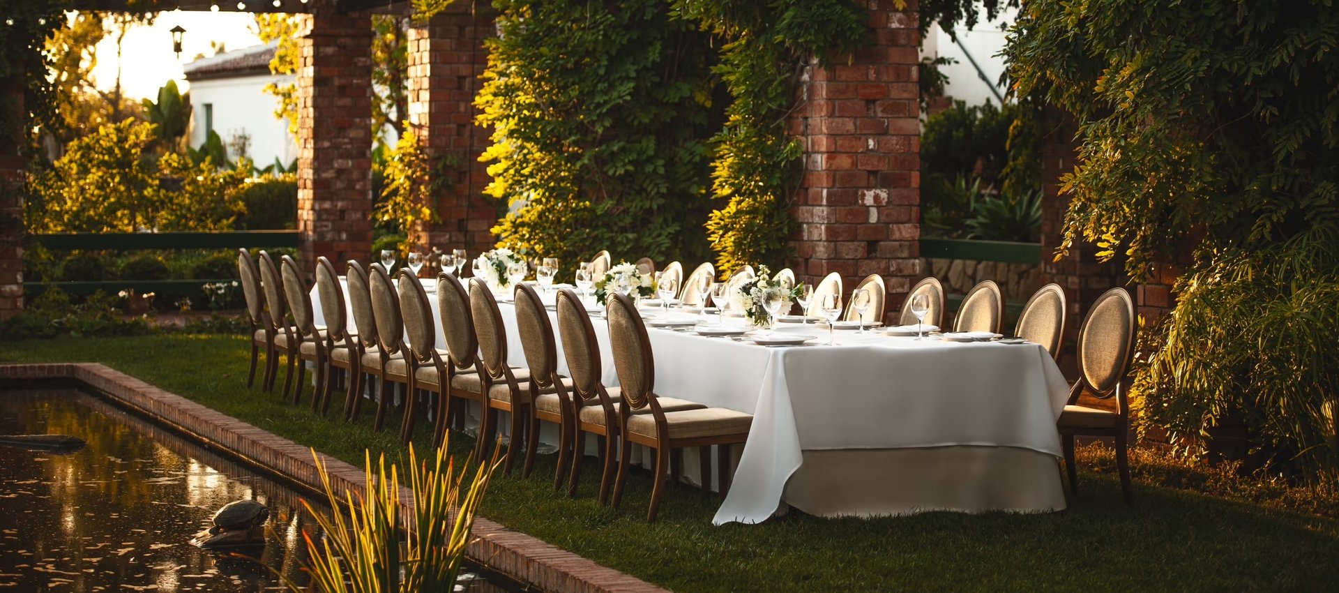 Elegant outdoor banquet table set for a wedding dinner in a lush garden with brick pillars.