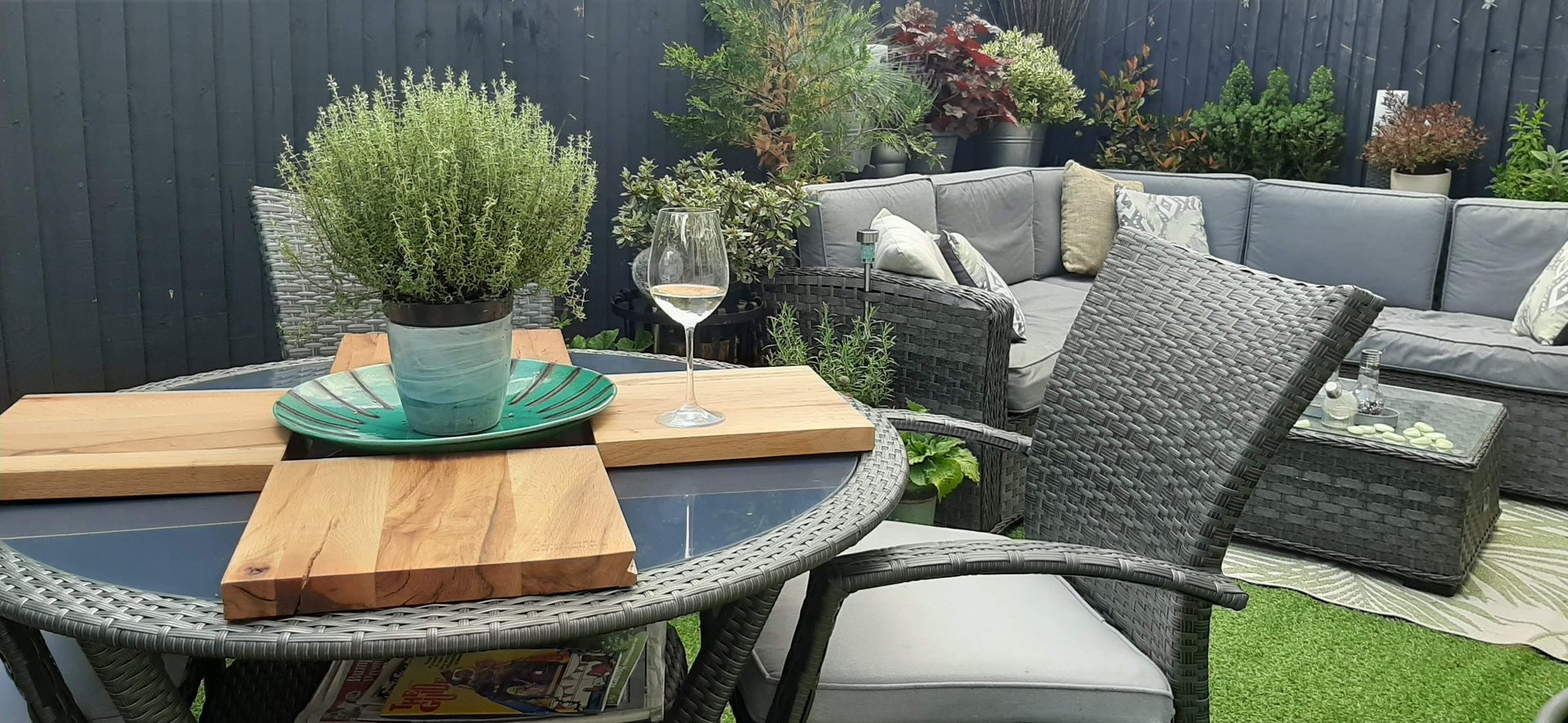 Modern garden patio with grey wicker furniture, a wine glass, and potted herbs on a glass table.
