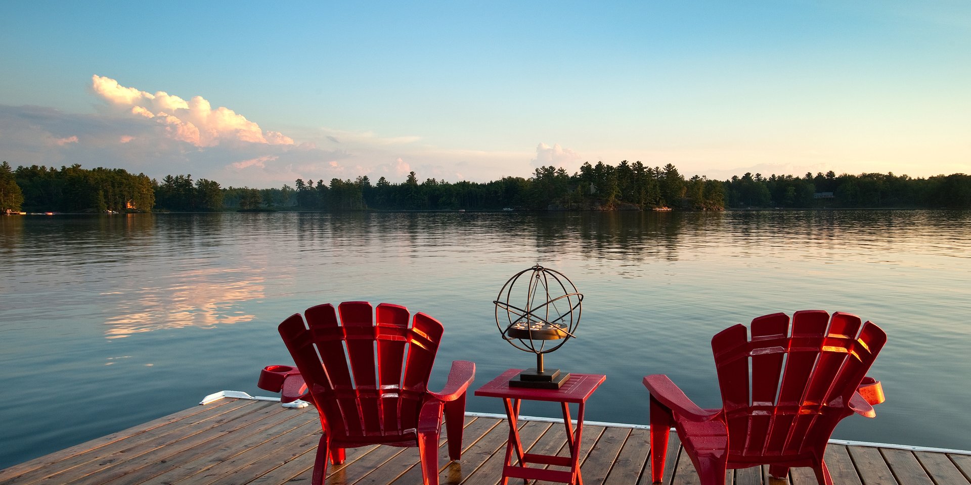 Red Adirondack chairs on a wooden lake dock at sunset with a scenic forest background.