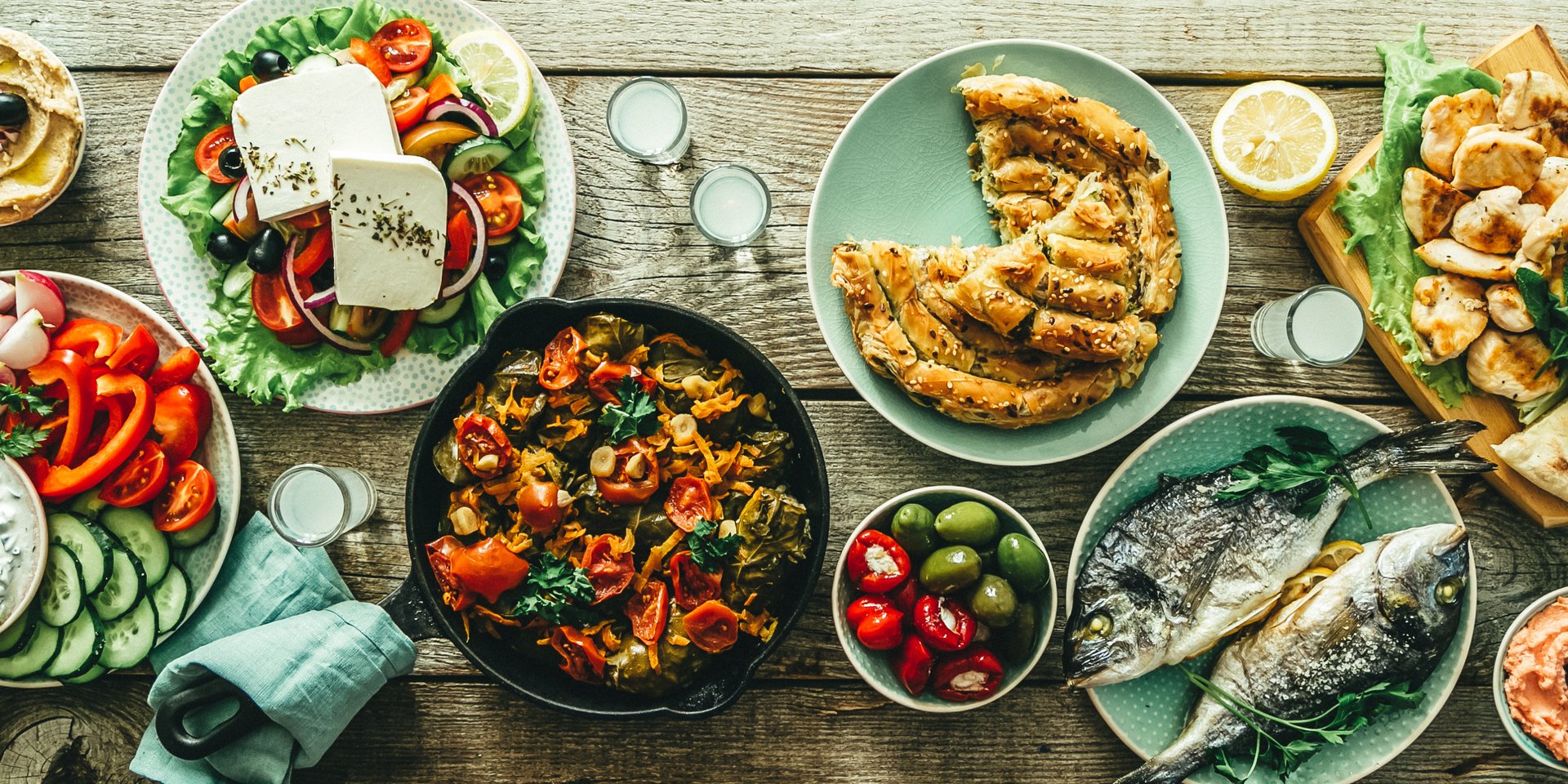 Traditional Greek food spread with grilled fish, feta salad, spanakopita, and olives on a rustic wooden table.