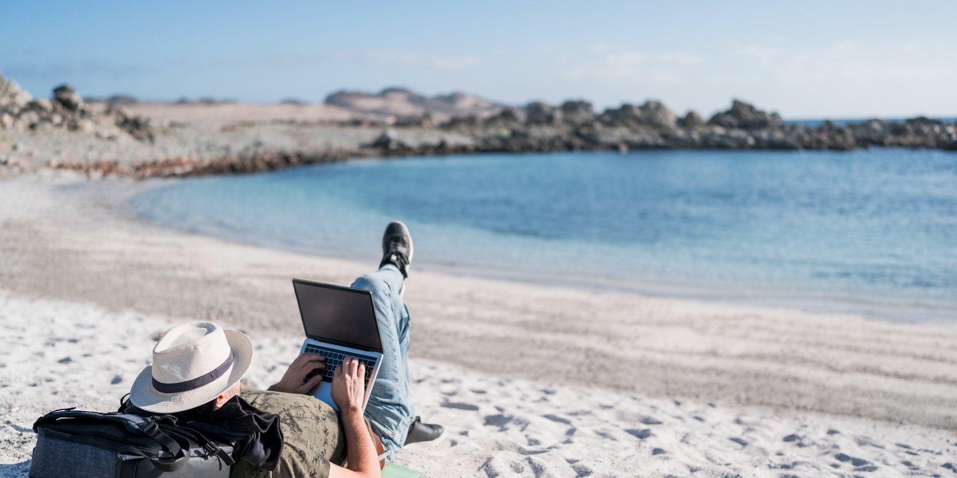 Digital nomad working on a laptop while relaxing on a white sand beach near the blue ocean.