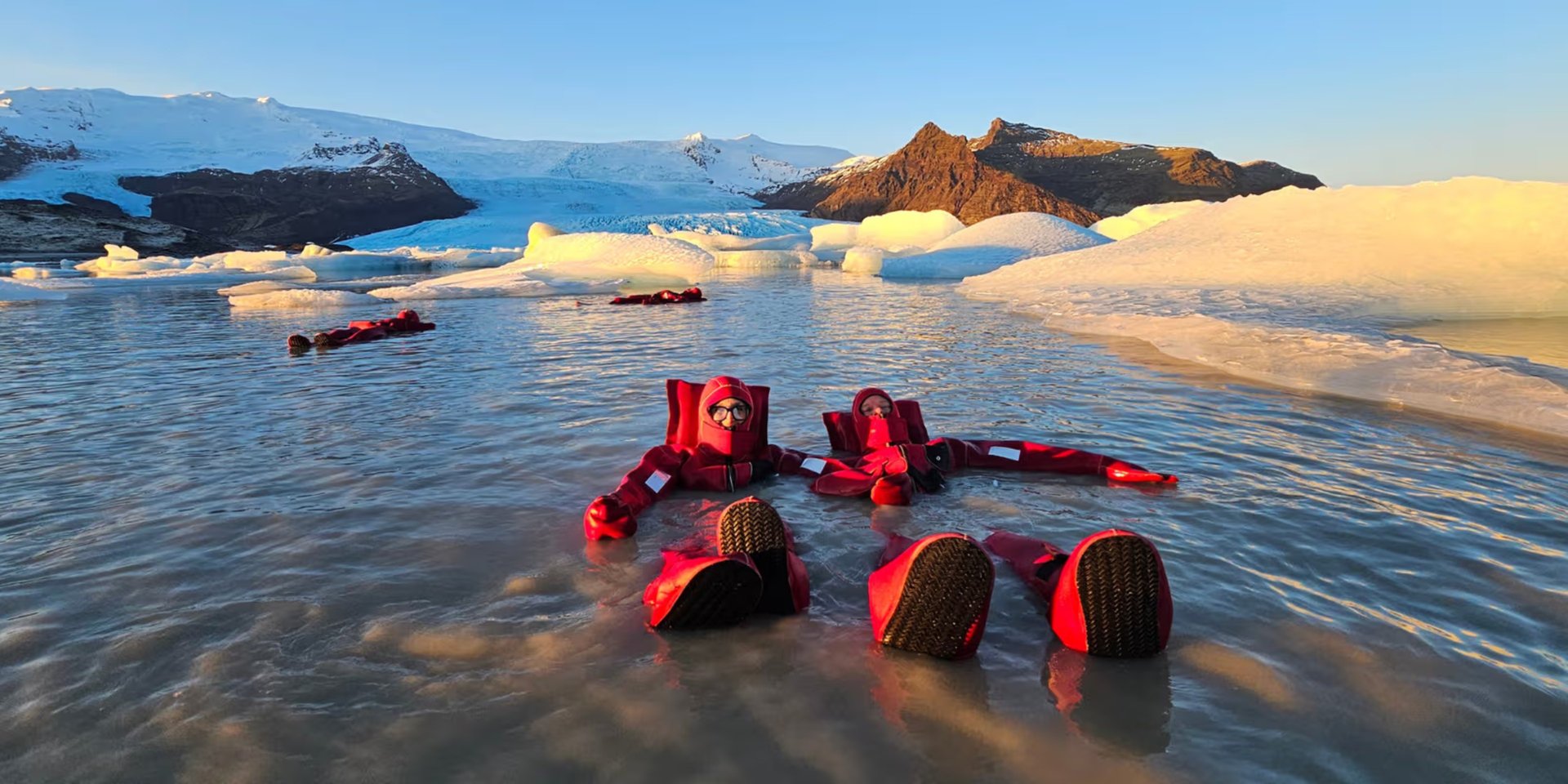Tourists in red dry suits floating in an Icelandic glacier lagoon surrounded by icebergs.
