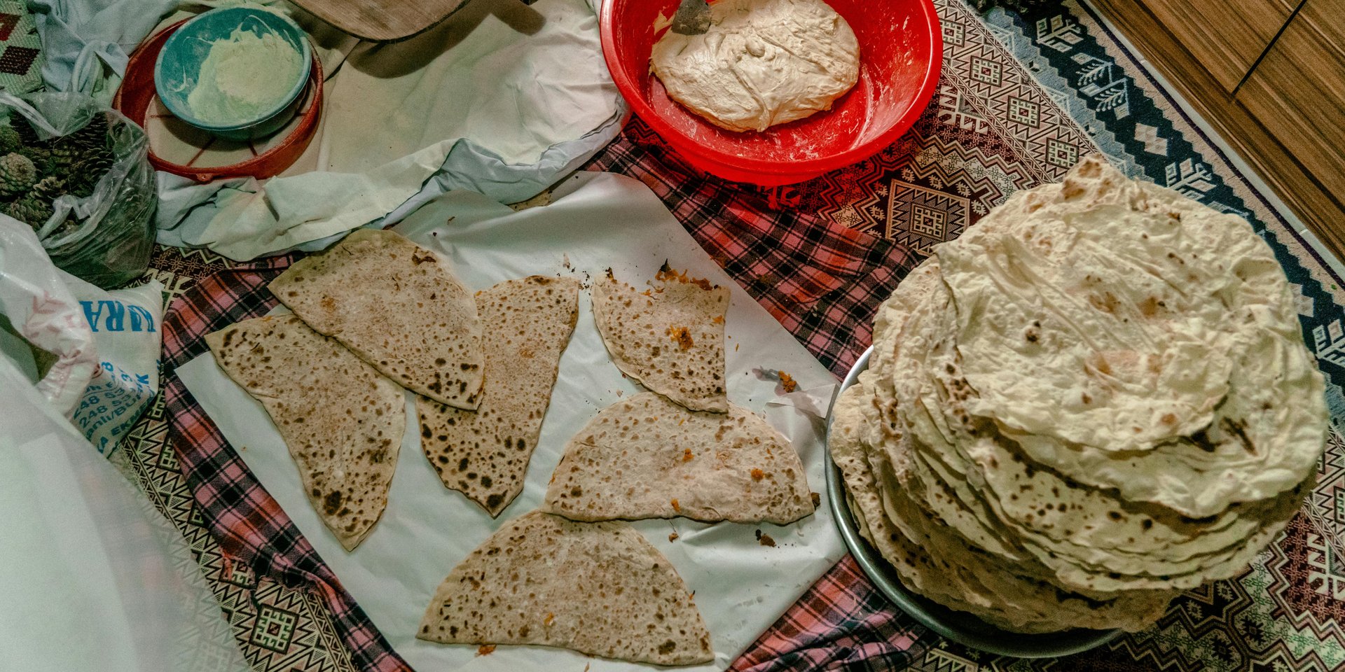 A stack of traditional flatbread and pieces of lavash next to a red bowl of fresh dough on a patterned rug.