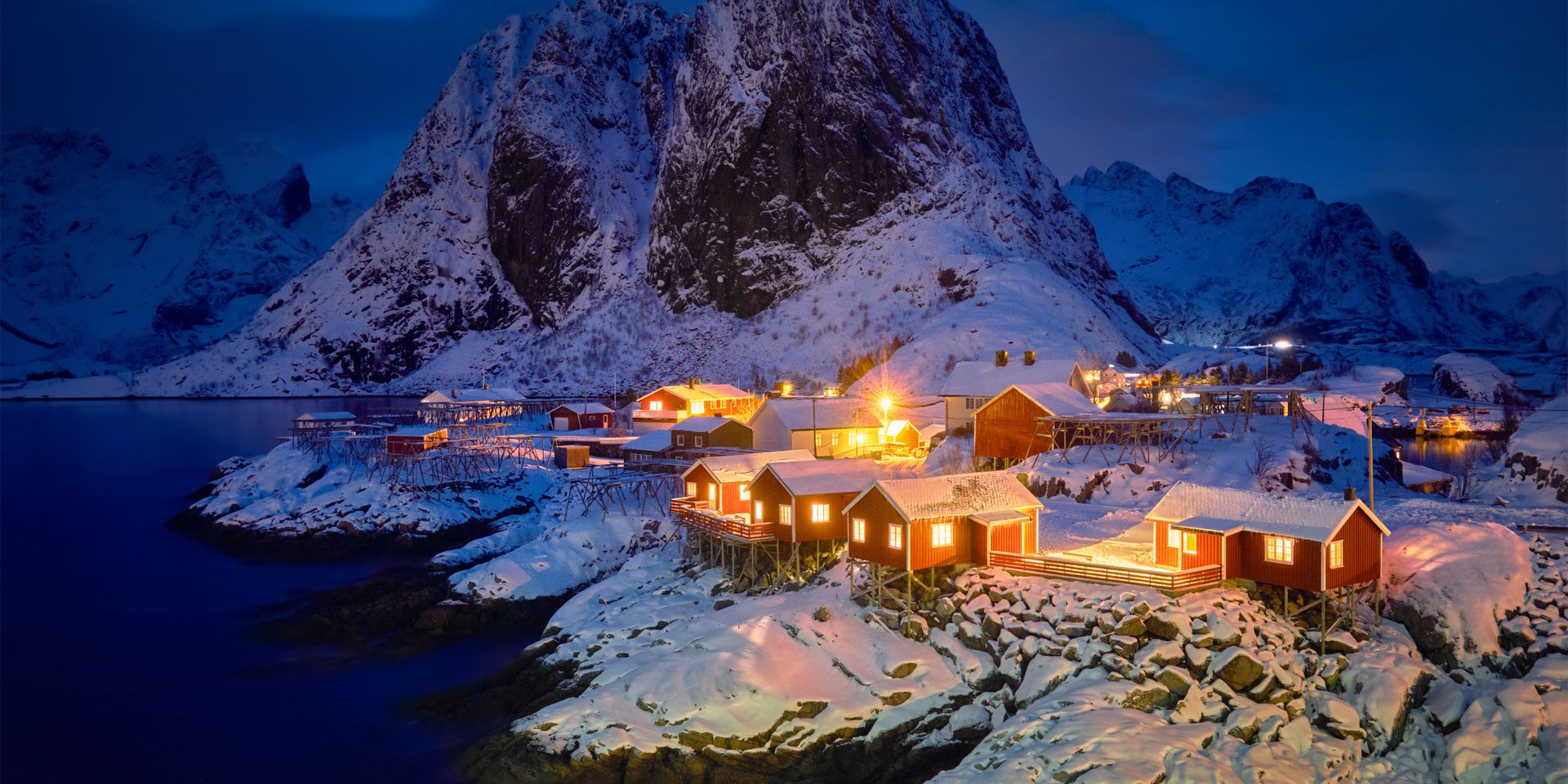 Illuminated red fishing cabins in Reine, Lofoten Islands, nestled under snow-capped mountains at twilight.
