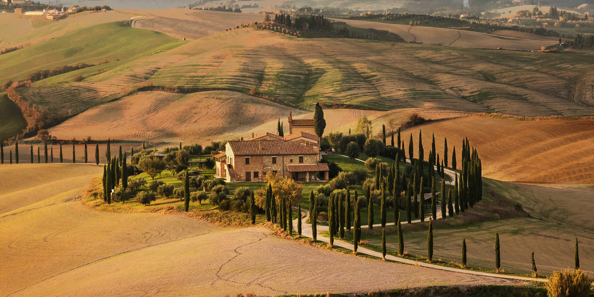 A scenic stone villa on a Tuscan hillside surrounded by cypress trees and rolling golden fields at sunset.