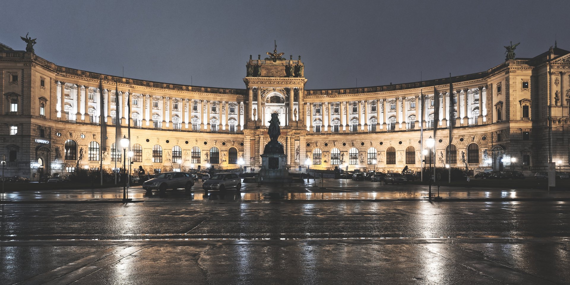 Illuminated facade of Hofburg Palace at night in Vienna, Austria, with wet pavement reflections.