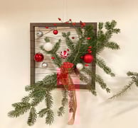 Decor de Crăciun în ramă de lemn, cu crenguțe de brad, globuri roșii și fundă festivă.