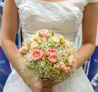 Buchet de mireasă cu trandafiri roz pal și gypsophila, ținut de o mireasă în rochie albă brodată.