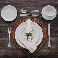 Mesa posta com sousplatt de rattan e jogo de louças de porcelana branca com talheres prateados.