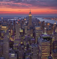 A beach scene with numerous blue beach cabanas and lounge chairs spread across the white sandy beach. A modern, curved high-rise building stands in the background, flanked by palm trees. The sky is clear and blue, creating a serene and inviting atmosphere.