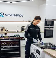 A female Novus-Pros technician in a gray and black uniform is meticulously repairing a washing machi