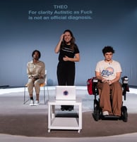 A black person, a white BSL signer, and a mixed-race electric wheelchair user sit on the stage