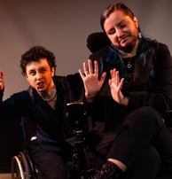 A mixed race manual wheelchair user and a white electric wheelchair user hold up their hands