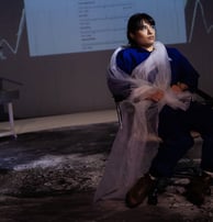 An Iranian woman in blue overalls sits, twisted in long white fabric