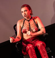 A white person in an electric wheelchair wearing red leather trousers, harness, and tape