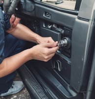 An assortment of locks and keys is displayed in a shop window. The window is framed with brown wood, and a wire mesh covers the glass. Various types of locks, including padlocks and antique keys, are visible. There are also licenses and signs visible inside the shop.