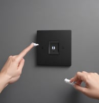 A dimly lit area with a dark background is visible. There is a central object resembling a security camera or electronic device. Below it, there is a rectangular white box with some graphics or text on its side. Cables or wires appear to be hanging or connected among the items.