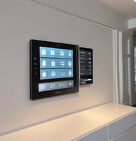 Two surveillance cameras are mounted on a stark, minimalist wall. The cameras have a teal body with orange accents and are connected by black cables. They are angled in different directions, suggesting active observation.