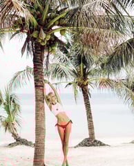 A blonde model wearing a white crop top, red bikini bottoms, and luxury jewelry poses against beach