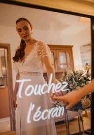 Une femme habillée en robe de mariée touche l'écran tactile du miroir photobooth de pixtoevent