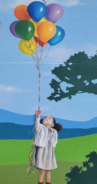 Fresque peinte sur façade. Une fillette sur un muret, tient une grappe de ballons colorés.