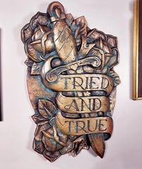 A plaque in the style of traditional tattoo art. Dagger with roses and a banner saying Tried & True