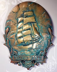 Image of a custom plaque. It shows a tall ship sailing. Below it is a scrollwork frame with a banner  that says Hold Fast. 