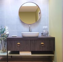 Salle de bains avec meuble en bois, poignées déco, tablette et miroir rond. vasque posée sur plan.