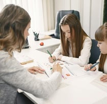 "Home tutor teaching two students, providing personalized instruction in a comfortable setting."