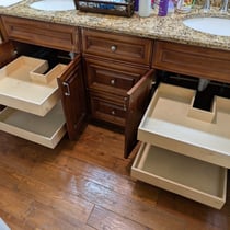 A bathroom vanity with four roll-out shelves installed. Two roll-outs are notched to fit around plumbing.