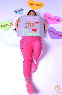 Nurse Nova holding a card that says "Trust the Process" covering her face, excluding her eyes, laying surrounded by hearts.