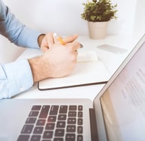 Homem escrevendo em um caderno, com laptop e celular sobre a mesa.