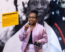 A woman in a pink jacket, pink and white skirt, pink earrings, and glasses holding a microphone