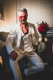 dapper older gentleman in white vintage 3 piece tuxedo in dim light sitting by red roses
