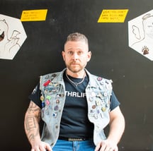 man in goatee with denim vest covered in pins with cartoon angel and demon on black background
