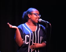 a Black woman in a navy baseball jersey and glasses speaking into a microphone