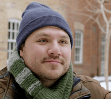 A young man in a scarf and toque smiles softly 