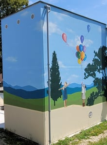Fresque peinte sur façades. Un paysage simplifié en arrière plan, un muret en trompe-l'oeil.