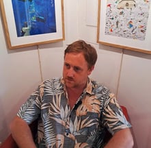 Artist seated in a red chair beneath framed digital prints of underwater and courthouse-themed artwo