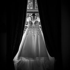  Das Schwarz-Weiß-Bild zeigt ein elegantes Hochzeitskleid mit Spitzenapplikationen, das auf einem Kleiderbügel vor einem Fens