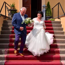 Das Bild zeigt eine lächelnde Braut in einem weißen Hochzeitskleid, die von einem Mann in einem blauen Anzug mit Krawatte ein
